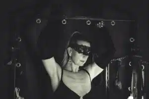 Lady Dawina Moon mit erhobenen Armen an einem schwarzen Gestell, Spitzenmaske und lange Handschuhe