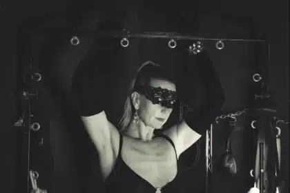 Lady Dawina Moon mit erhobenen Armen an einem schwarzen Gestell, Spitzenmaske und lange Handschuhe